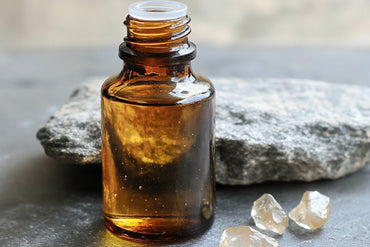 A small amber glass bottle of magnesium oil sits on a grey stone surface next to a larger smooth rock and two clear crystals, highlighting the natural and mineral-rich properties of the product.